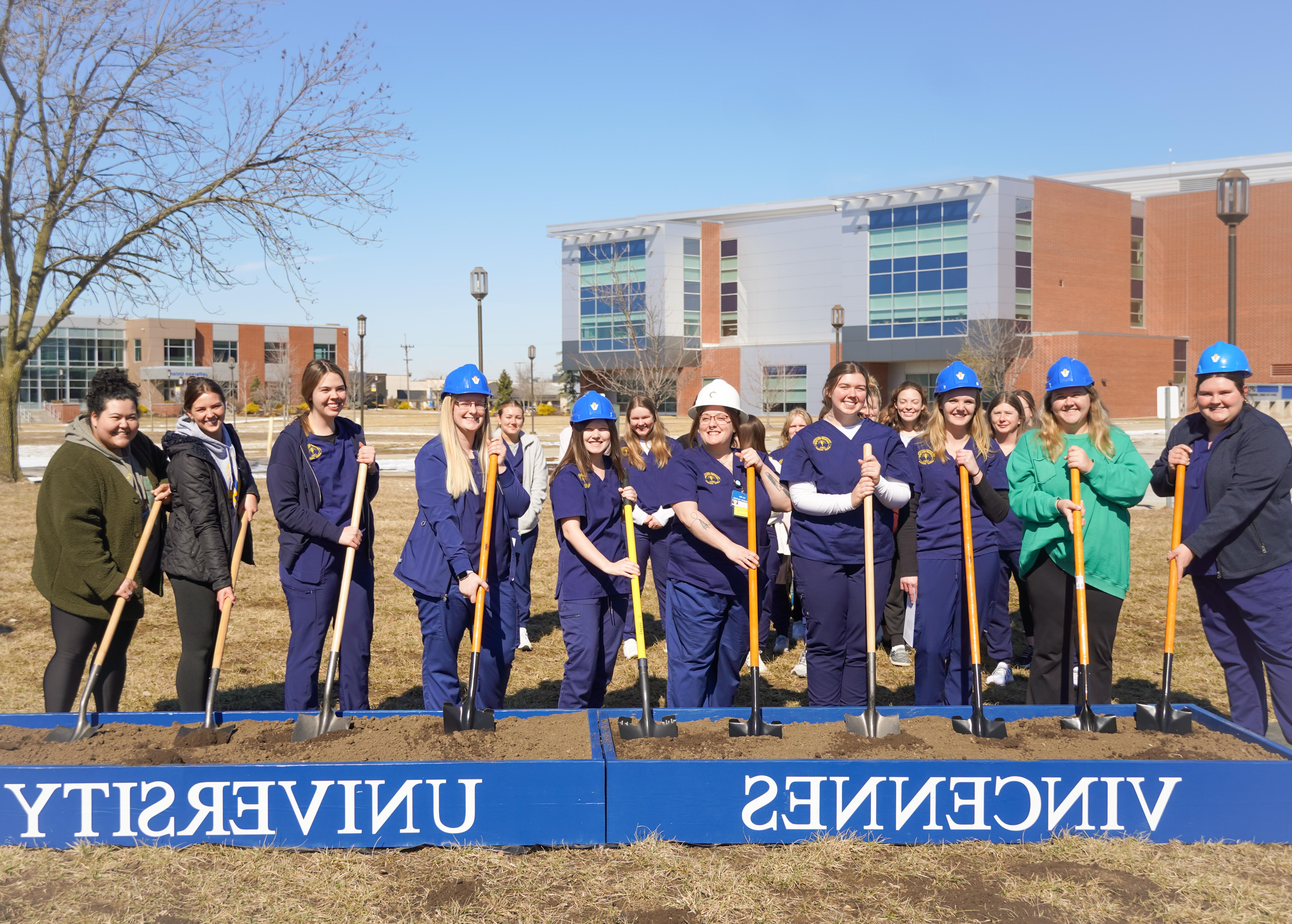 Female students wearing hard hats and scrubs while holding shovels inside boxes filled with dirt that have Vincennes University printed on them.